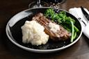 A plate with a grilled steak topped with butter, served with mashed potatoes and steamed broccolini. Salt is sprinkled on top. A knife, fork, and napkin are beside the plate on a dark wooden table.