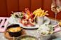 A seafood meal on a table features half of a cooked lobster with fries in a metal cup, a small salad, and a glass of white wine. Accompaniments include a baked dish covered with cheese and a bowl of leafy greens. Utensils and napkins are placed nearby.