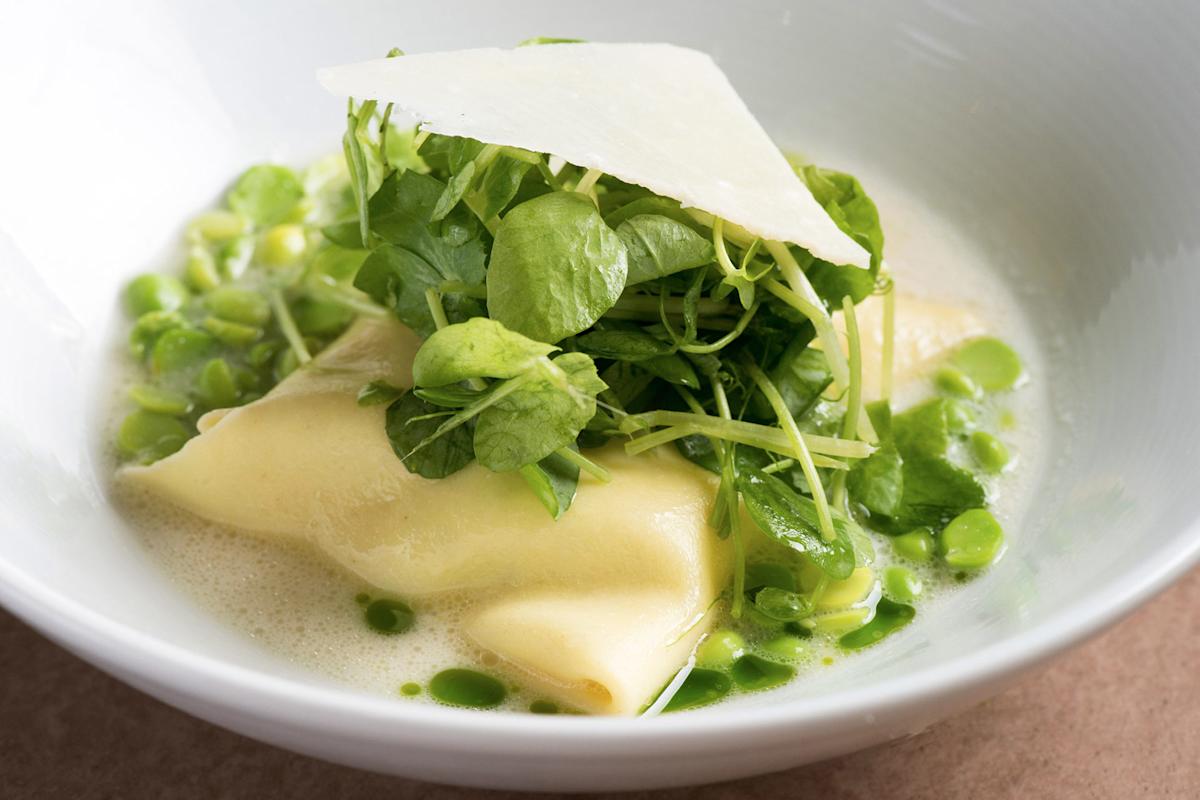A white bowl containing ravioli topped with fresh green peas, pea shoots, and a thin slice of cheese, all sitting in a light green broth with small droplets of oil.