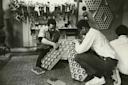 Two people kneel on the floor, assembling a structure made of large clear bulbs in a workshop filled with tools and pegboards. Another person sits nearby with their back to the camera.