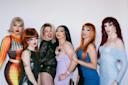 Six drag queens pose together in colorful, glamorous outfits with bold makeup, wigs in various colors, and confident expressions against a plain white background.