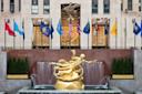 A golden statue of Prometheus stands in front of a fountain at Rockefeller Center, with national and state flags displayed above and a building entrance in the background.
