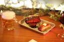 A tray with glazed ribs on lettuce and a slice of cornbread with butter sits on a wooden table. A glass mug of beer is on the left, and a glass of red wine is on the right. Greenery decorates the backdrop.