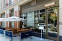 Street view of a restaurant called Mishik with large glass windows, wooden outdoor tables and chairs under white umbrellas, and a menu stand by the entrance. Blue barriers separate the patio from the sidewalk.