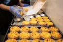 A person wearing blue gloves is placing baked Portuguese egg tarts into a white box, with many more egg tarts arranged on trays in the foreground.