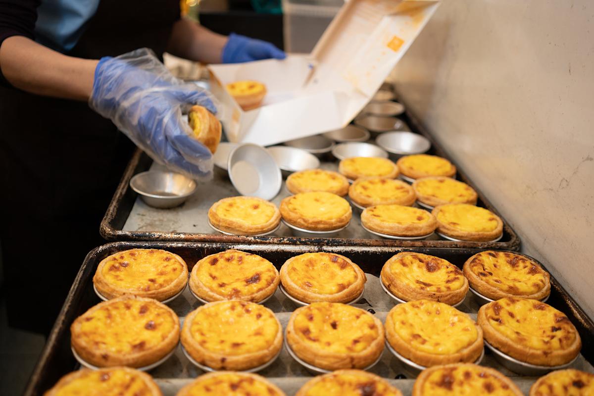 A person wearing blue gloves is placing baked Portuguese egg tarts into a white box, with many more egg tarts arranged on trays in the foreground.