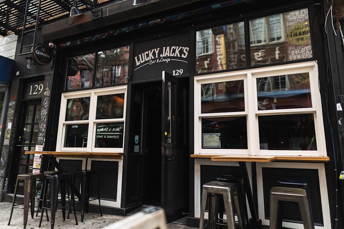 Street view of Lucky Jack’s Bar & Lounge with large windows, bar stools, a black exterior, and address number 129 displayed above the entrance. Reflections of nearby buildings are visible in the windows.