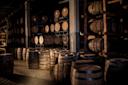 Dimly lit storage room filled with wooden barrels stacked on racks and arranged on the floor, creating a rustic atmosphere often associated with distilleries or wineries.