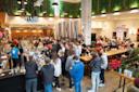 A busy brewery with people gathered around the bar and seated at tables. Large steel tanks are visible in the background. The room is decorated with greenery and bright lights, creating a lively atmosphere.