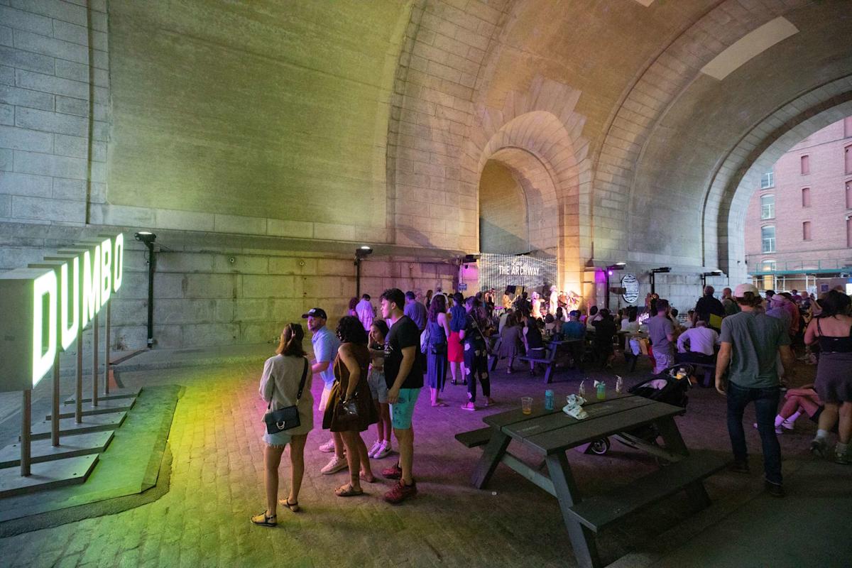 A crowd gathers under a large stone archway at night, near a glowing "DUMBO" sign, watching a live band perform on a stage; picnic tables and colorful lighting are visible.