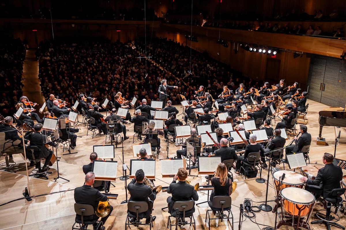 A symphony orchestra performs on stage in a large concert hall, with a conductor leading musicians playing strings, woodwinds, brass, and percussion. The audience is seated in the background.