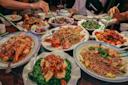A table filled with various Chinese dishes, including noodles, fish, stir-fried vegetables, and meat, surrounded by people using chopsticks to serve themselves. Bottles and teacups are also on the table.
