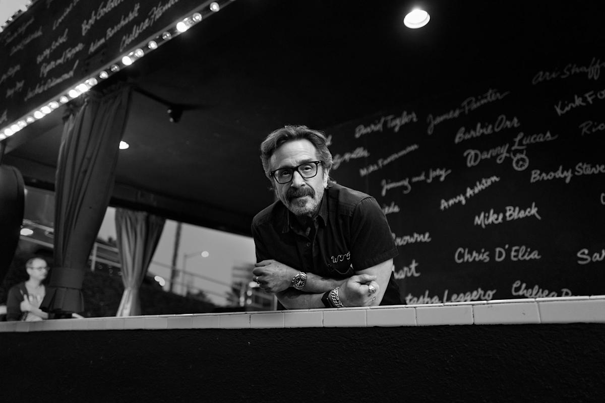 A man with glasses leans forward on a ledge, looking at the camera in a black-and-white setting. The wall behind him is covered in handwritten names. A person sits in the background on the left.