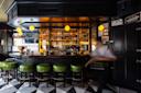 A stylish bar with green stools, a black counter, shelves of liquor bottles, and four hanging yellow globe lights. A blurred figure moves past on a floor with a black and white geometric pattern.
