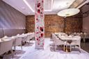 Modern restaurant interior with white marble floors, white chairs and tables set for dining, a decorative column with red and black floral art, textured walls, and a large round hanging light fixture.