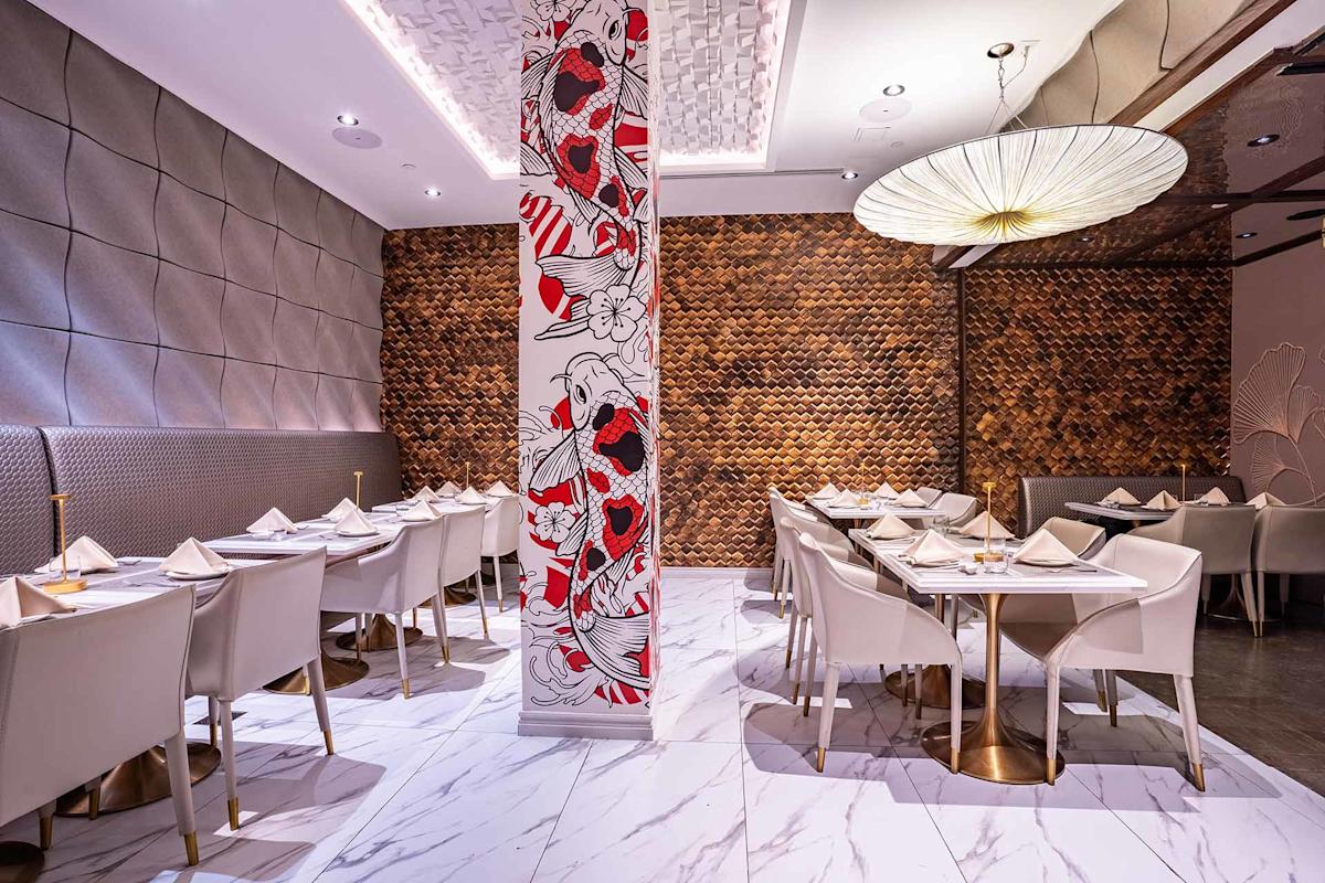 Modern restaurant interior with white marble floors, white chairs and tables set for dining, a decorative column with red and black floral art, textured walls, and a large round hanging light fixture.