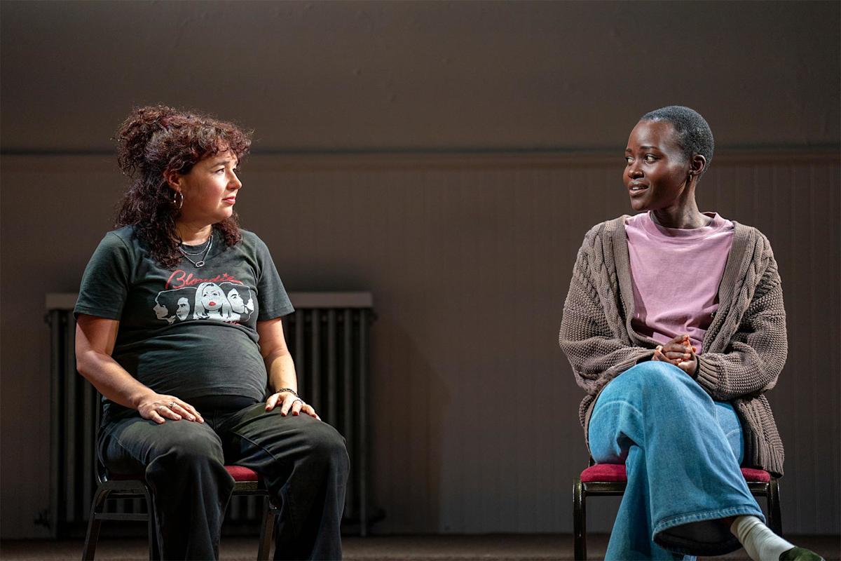 Two women sit on chairs facing each other, engaged in conversation. One woman is pregnant and wears a black graphic T-shirt. The other wears a mauve shirt and cardigan. Both appear serious and focused.