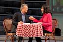 A man and woman sit across from each other at a table with a red and white checkered tablecloth. They are holding hands, smiling, and appear engaged in a conversation. The background features shelves filled with books.
