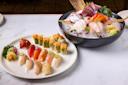 A plate of assorted sushi and sashimi with a variety of colorful fish, garnished with herbs and vegetables, is displayed on a marble table. Next to it, a bowl filled with ice holds more sashimi and decorative garnishes.