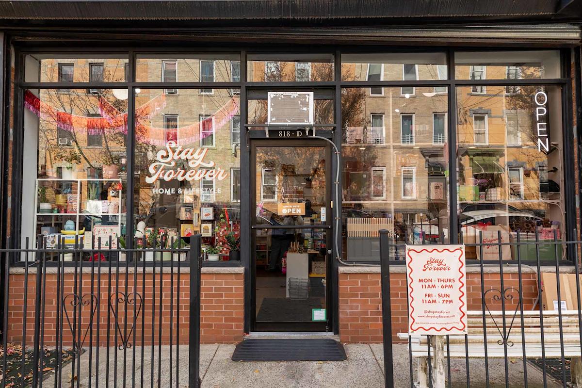 Storefront view of “Stay Forever Home & Gifts” with large windows, colorful decorations inside, and an “OPEN” sign. A sandwich board outside lists store hours. Residential buildings are reflected in the glass.