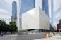 A large, modern, white cube-shaped building with a textured surface stands at a city intersection, surrounded by tall glass skyscrapers; pedestrians cross the street and trees line the sidewalk.