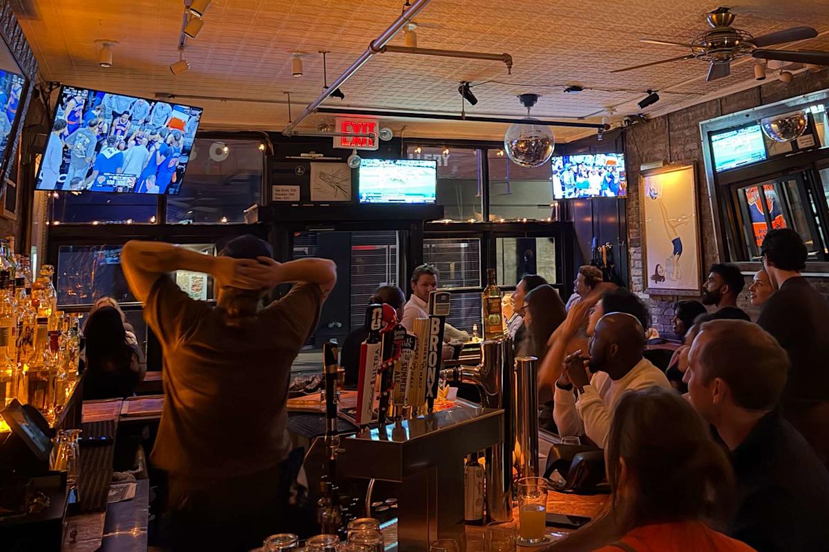 A lively bar with people gathered around watching a sports game on TV screens. The bar is warmly lit, with drinks on the counter and various beer taps visible. Some patrons are cheering or intently watching the game.