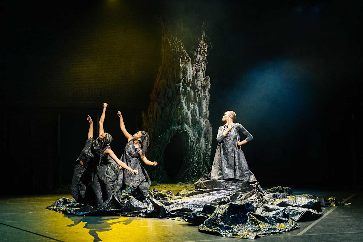Three dancers in dramatic black costumes perform on stage with expressive poses. A large, tree-like structure and dark, artistic lighting create a mystical, theatrical atmosphere.