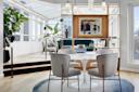 A modern dining area with four gray chairs around a white marble tulip table, set on a circular rug. Behind, a gold-framed divider with art separates the dining and living areas, with large windows and indoor plants throughout.
