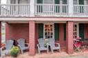 Four gray Adirondack chairs sit on a brick patio outside a rustic building with red brick walls, green shutters, and potted yellow and white flowers. A small table with red chairs is on the right.