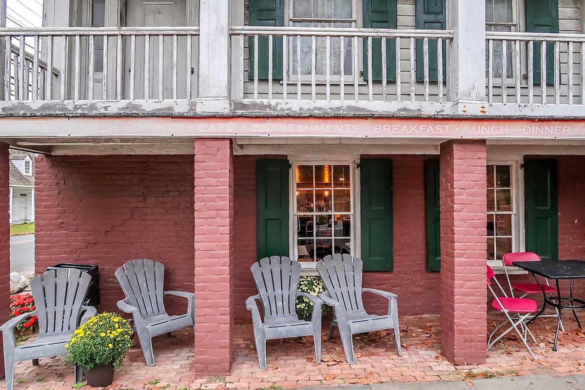 Four gray Adirondack chairs sit on a brick patio outside a rustic building with red brick walls, green shutters, and potted yellow and white flowers. A small table with red chairs is on the right.