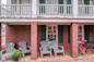 Four gray Adirondack chairs sit on a brick patio outside a rustic building with red brick walls, green shutters, and potted yellow and white flowers. A small table with red chairs is on the right.