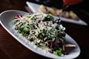 A close-up of a fresh green salad served on a white oval plate, topped with grated cheese, featuring leafy greens and thinly sliced vegetables, on a dark wooden table.