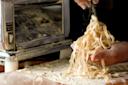 A close-up of hands holding freshly cut pasta noodles on a floured surface, with a metal pasta machine in the background and flour being sprinkled over the noodles.