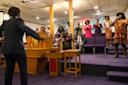 A choir sings enthusiastically on stage during a church service. A person stands below, arms outstretched, leading the choir. Congregants are seated watching the performance. The setting is decorated with warm tones and wooden elements.