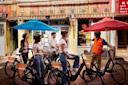 Five people wearing helmets stand with bicycles under colorful umbrellas on a street in front of a building with Chinese signage. One person in a reflective vest appears to be guiding or instructing the group.