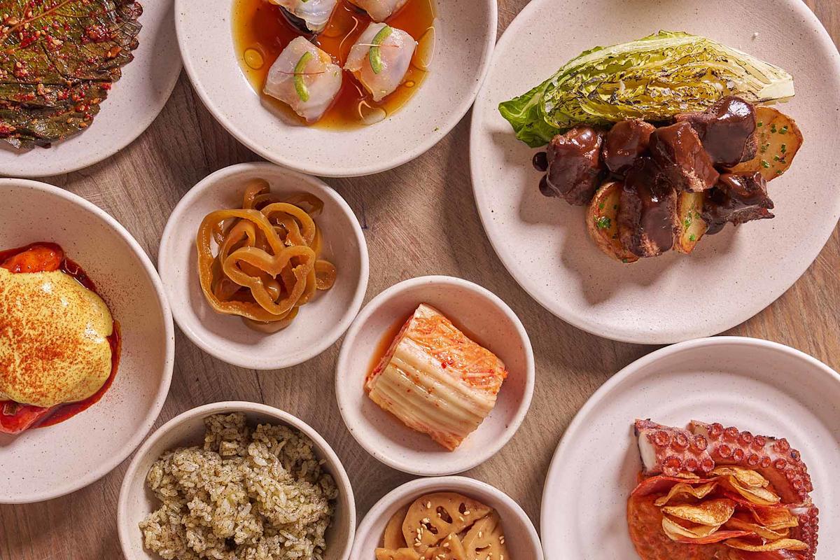 An overhead view of assorted small dishes, including grilled vegetables, sliced meats, seafood, pickles, kimchi, rice, and plates with savory sauces, all arranged on a wooden table.
