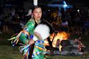 A person in traditional, colorful attire dances near a bonfire at night. They hold a fan made of feathers, and the background is filled with onlookers seated in a dimly lit area.