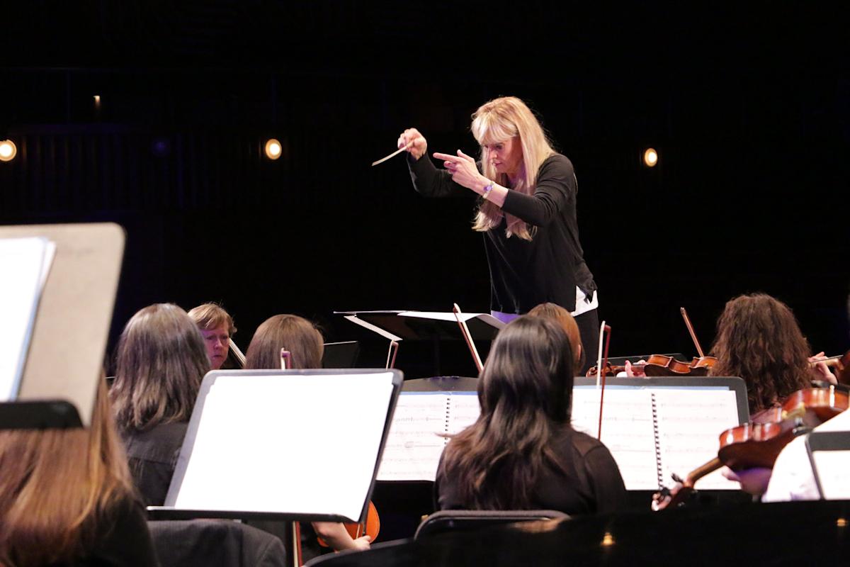 A conductor leads an orchestra, holding a baton high in one hand. Musicians seated in front of her focus on their sheet music and play string instruments. The scene is dimly lit, emphasizing the musicians and their music stands.