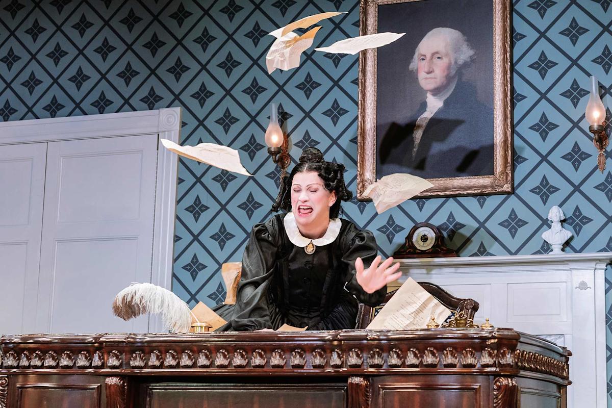 A woman in a black Victorian dress sits at a large wooden desk, looking frustrated as papers fly around her. Behind her, a portrait of George Washington hangs on a patterned wall.