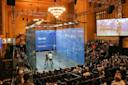 A squash match takes place in a glass court surrounded by a seated audience in a large, elegant hall with chandeliers. A large screen displays the match above the crowd.