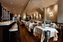 A stylish, elegant restaurant interior with neatly set tables, white tablecloths, wooden chairs, a long bar with stools, wine bottles, and warm ambient lighting. Framed art decorates the cream-colored walls.
