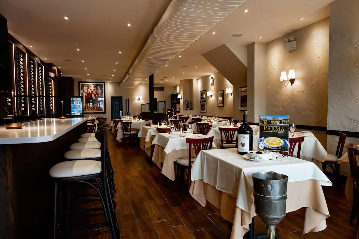 A stylish, elegant restaurant interior with neatly set tables, white tablecloths, wooden chairs, a long bar with stools, wine bottles, and warm ambient lighting. Framed art decorates the cream-colored walls.