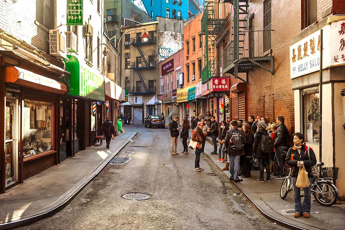 A group of people stand and walk along a narrow street lined with shops, signs, and buildings in a busy urban neighborhood, likely Chinatown, on a sunny day.