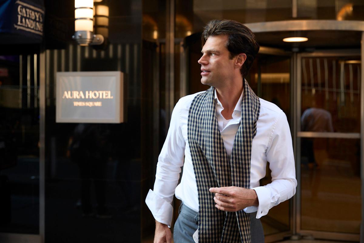 A man in a white shirt and checkered scarf stands in front of a hotel entrance, looking to the side. The hotel sign reads "Aura Hotel" and there's blurred background lighting.