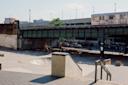 Two people are skateboarding under a green bridge at an urban skatepark as a subway train passes overhead. Concrete ramps and rails are visible, with city buildings in the background.