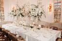 Elegant dining table set for a formal event, decorated with tall glass vases of white flowers and greenery, surrounded by beige chairs, white linens, and softly lit candles in a luxurious room.