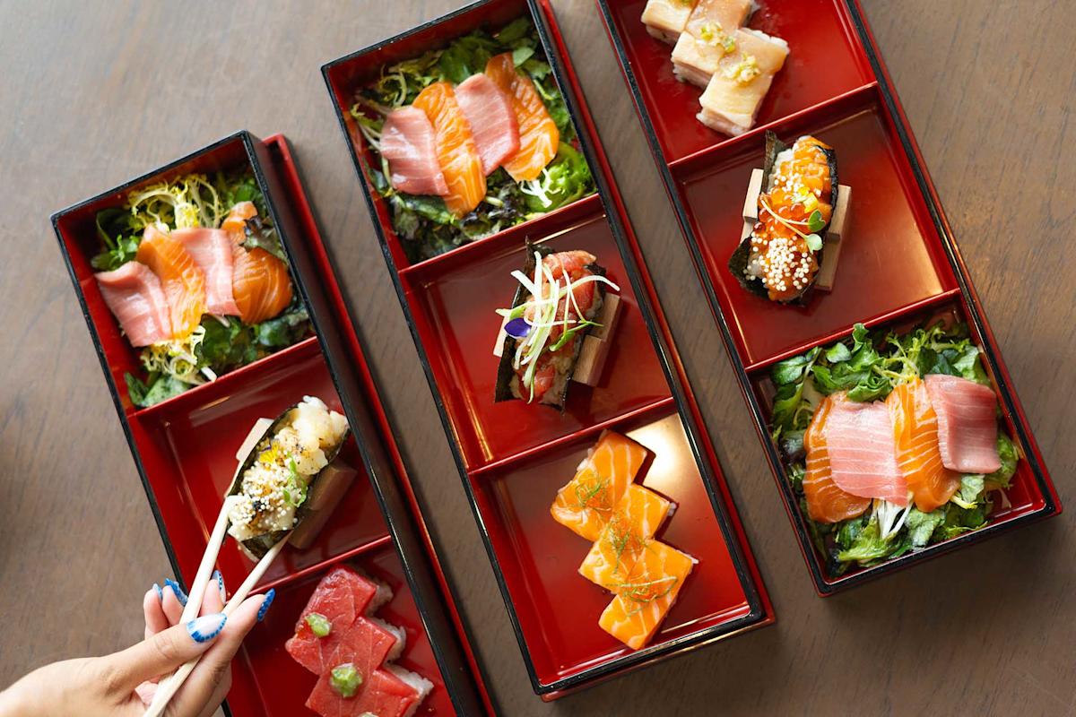 Four red bento boxes filled with assorted sashimi, sushi, and salads are arranged on a wooden table. A hand uses chopsticks to pick up a sushi piece from one of the boxes.