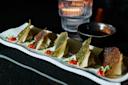 A row of gyoza with crispy tops is arranged on a rectangular plate, garnished with small red chili peppers and green herbs. A small bowl of dipping sauce and a lit candle are in the blurred background on a dark surface.