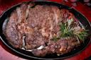 A cooked, seasoned steak sits on a black platter garnished with sprigs of fresh rosemary. The steak appears juicy with charred edges, set against a red background.