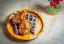 A wooden plate with two pieces of fried chicken on top of two dark purple waffles, drizzled with syrup, sits on a marble surface next to a small vase with pink flowers.